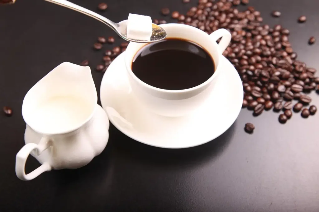 Tasse de café noir avec un morceau de sucre sur une cuillère et un petit pot de lait, grains de café autour