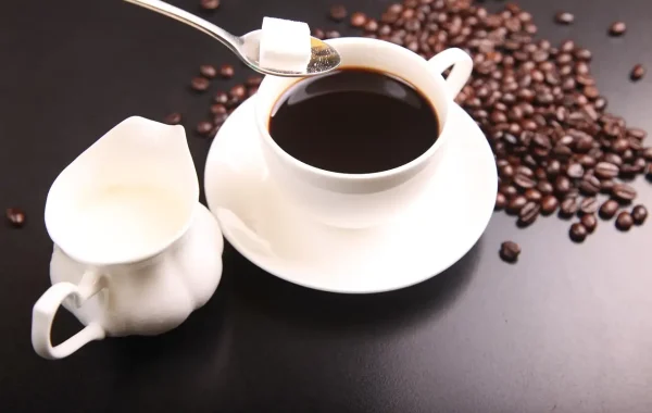 Tasse de café noir avec un morceau de sucre sur une cuillère et un petit pot de lait, grains de café autour