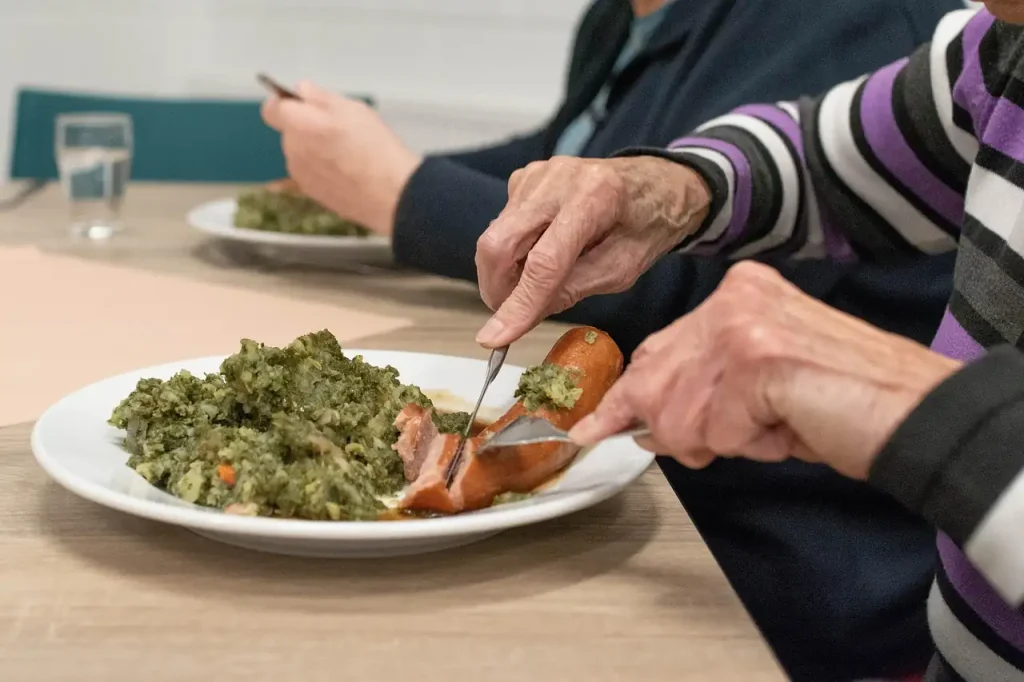 Mains d'une personne âgée coupant une saucisse dans une assiette de chou vert, à table