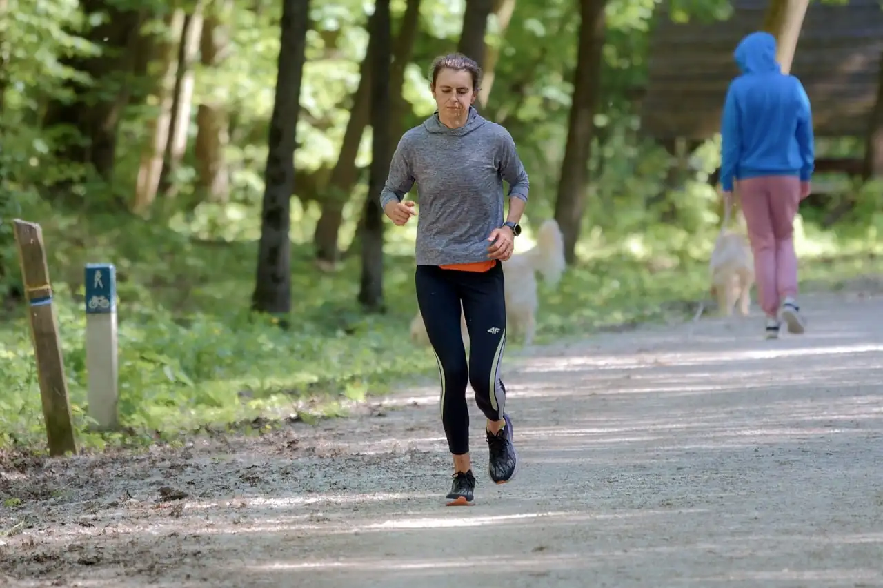 Femme en tenue de sport qui court sur un chemin de forêt, une montre au poignet, une promeneuse et son chien derrière elle