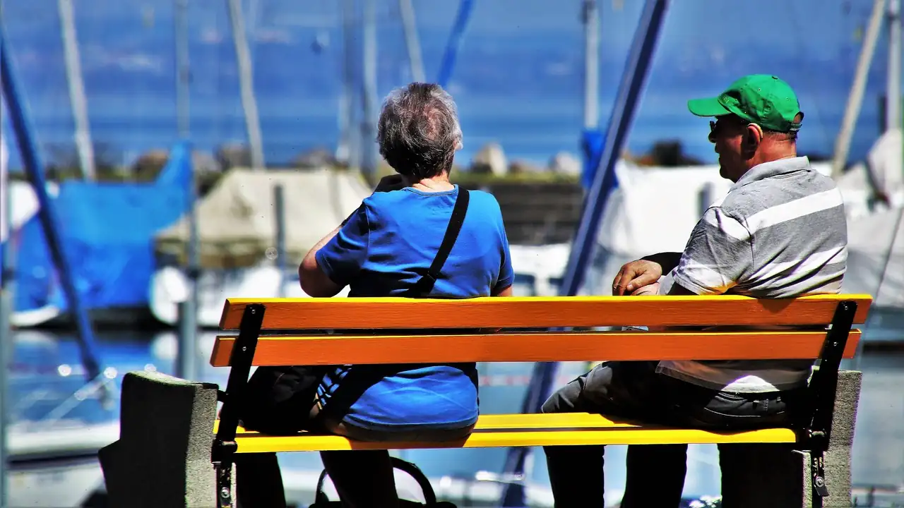 Couple âgé assis de dos sur un banc jaune face à un port de plaisance