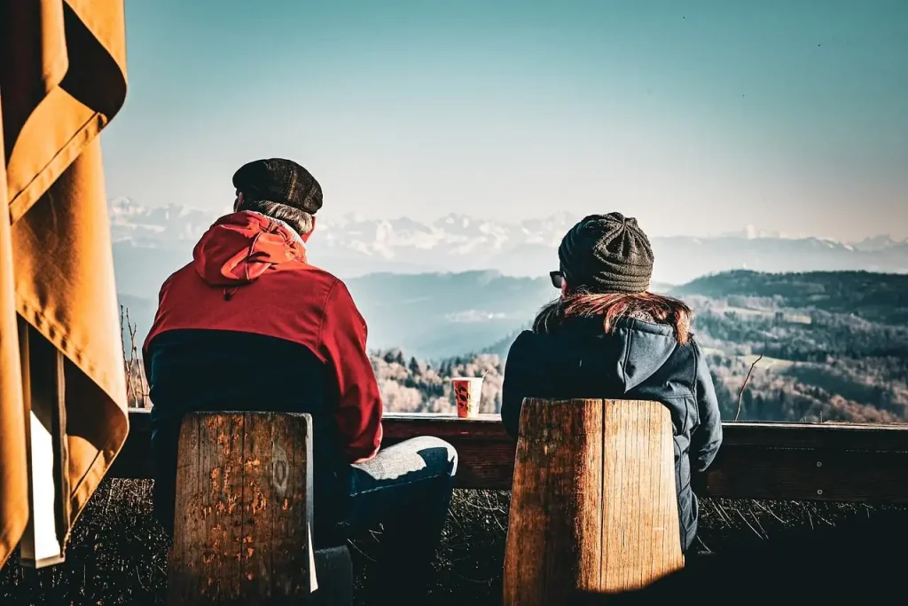 Deux personnes assises de dos sur des tabourets en bois devant un panorama de montagnes enneigées