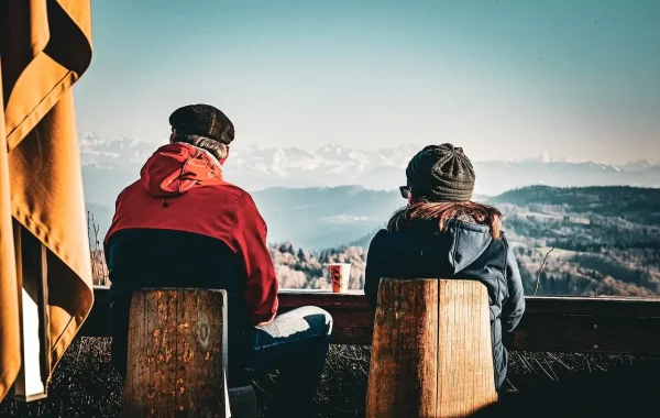 Deux personnes assises de dos sur des tabourets en bois devant un panorama de montagnes enneigées