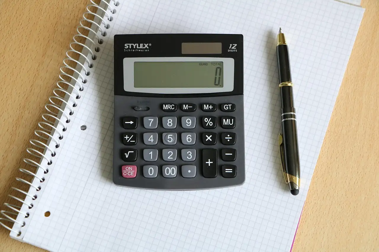 Calculatrice de bureau affichant zéro, posée sur un carnet à spirale quadrillé, à côté d'un stylo noir
