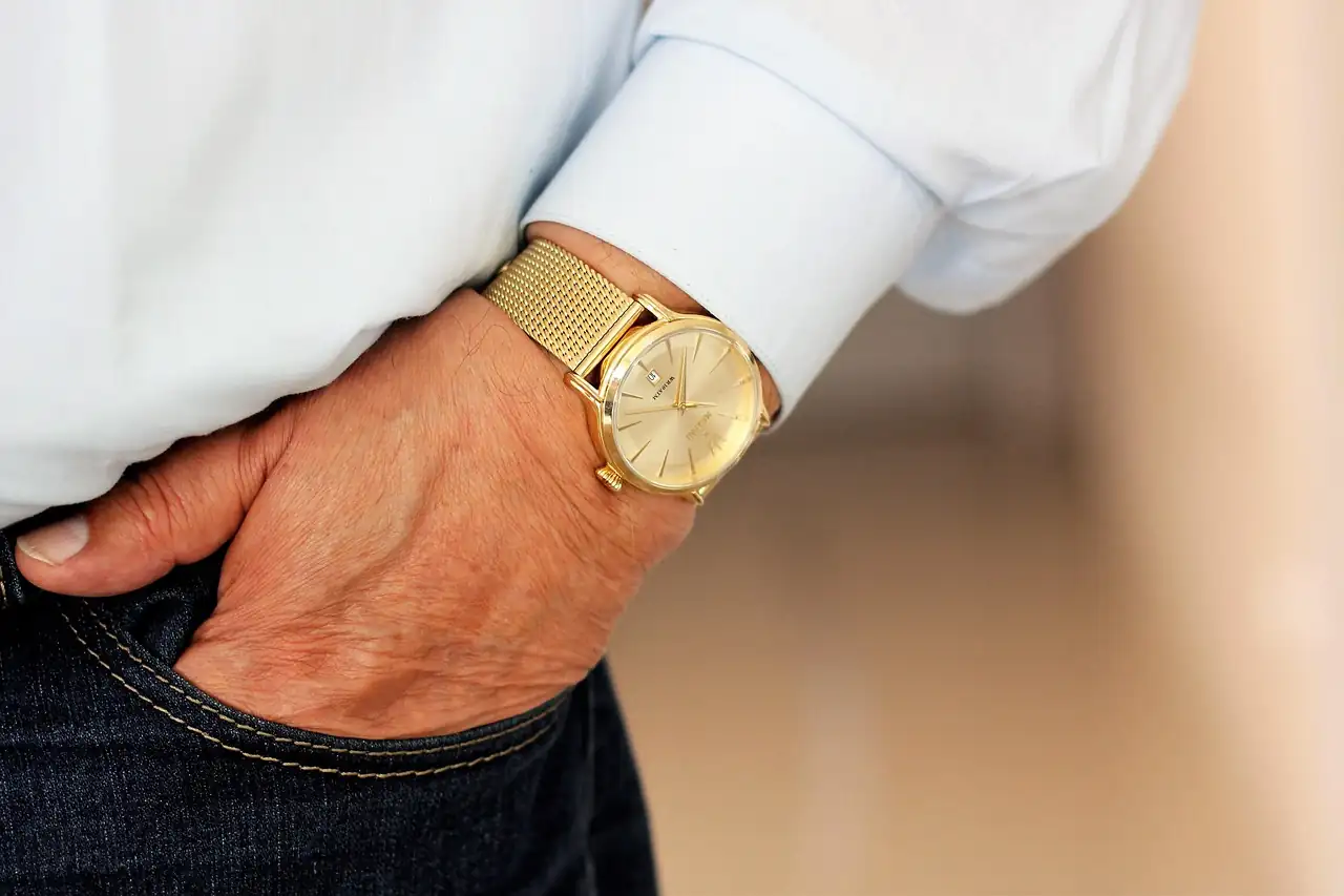 Poignet d'un homme en chemise claire portant une montre dorée à bracelet maille, main dans la poche