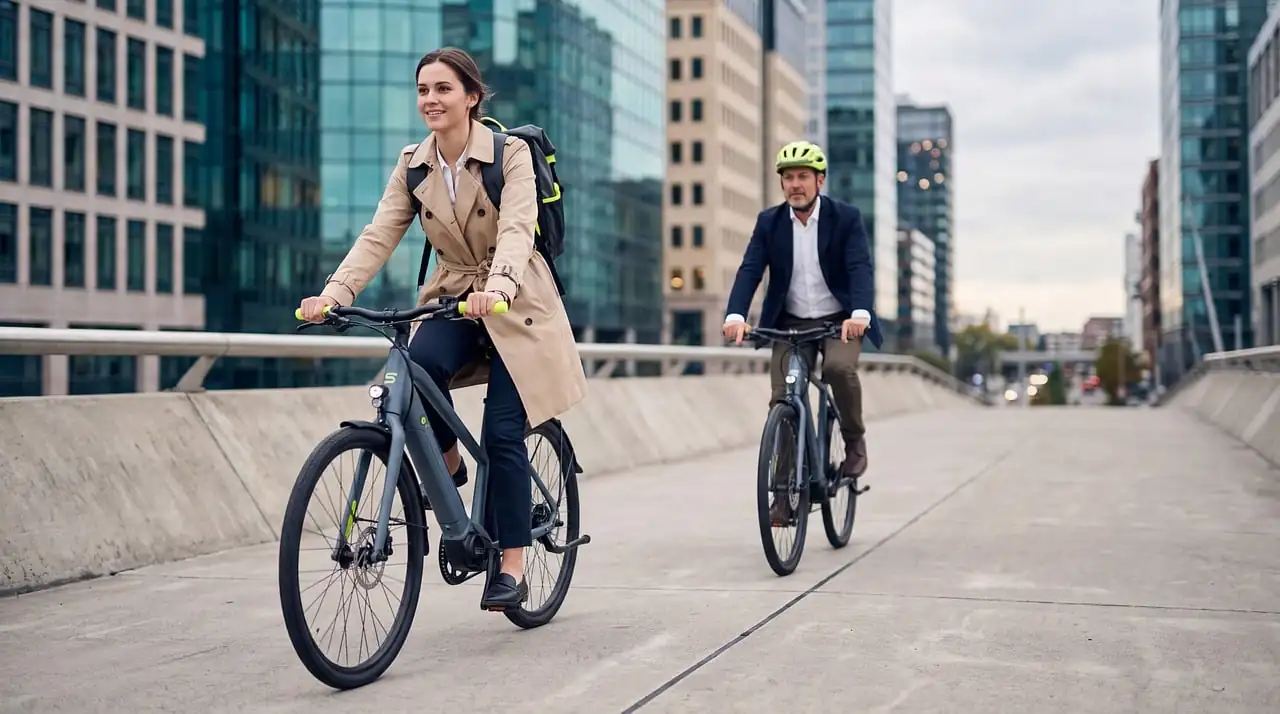 Femme en trench et homme en veste à vélo sur un pont, immeubles de bureaux en verre derrière eux