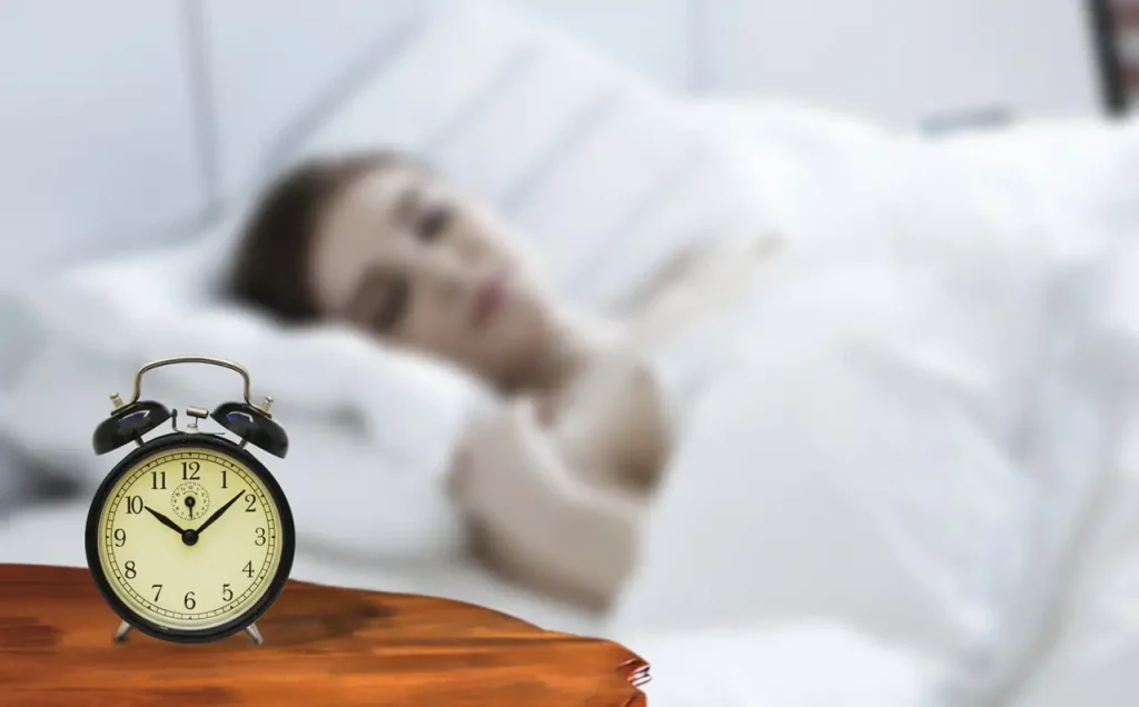 Réveil à cloches posé sur une table de nuit en bois, personne endormie floue dans un lit blanc