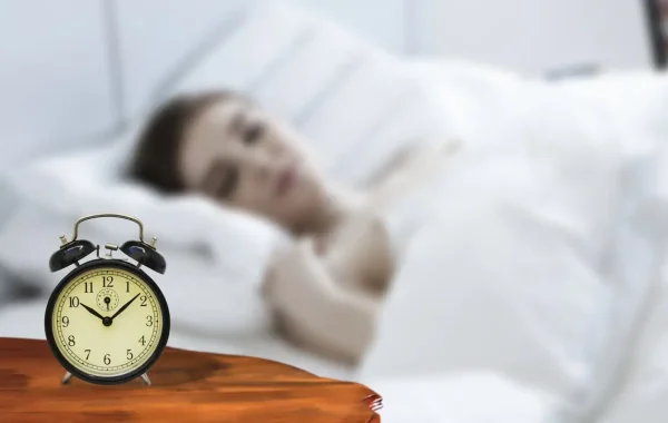 Réveil à cloches posé sur une table de nuit en bois, personne endormie floue dans un lit blanc