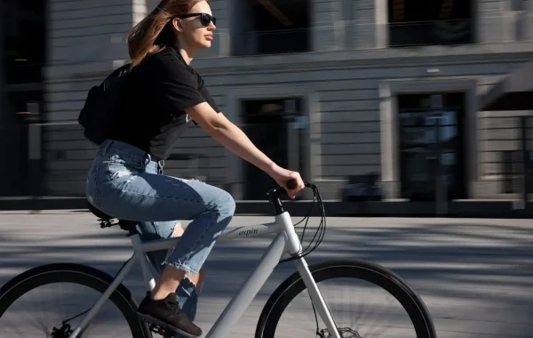 Jeune femme aux lunettes de soleil qui pédale sur un vélo blanc devant un immeuble de bureaux