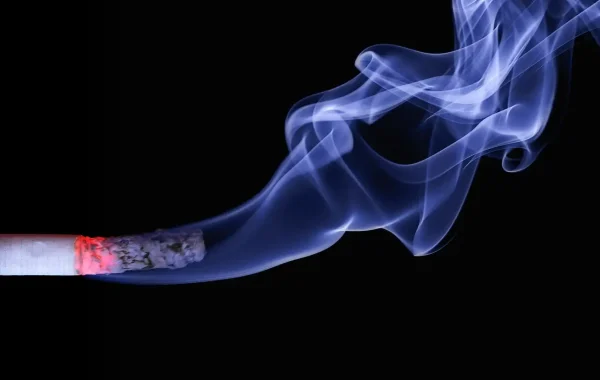 Cigarette allumée dont la fumée bleutée s'élève sur un fond noir