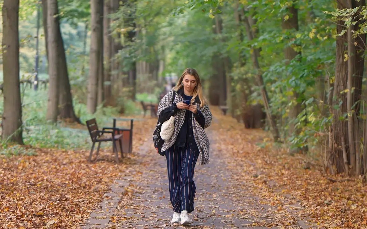 Jeune femme en manteau à carreaux qui marche dans une allée de parc couverte de feuilles mortes