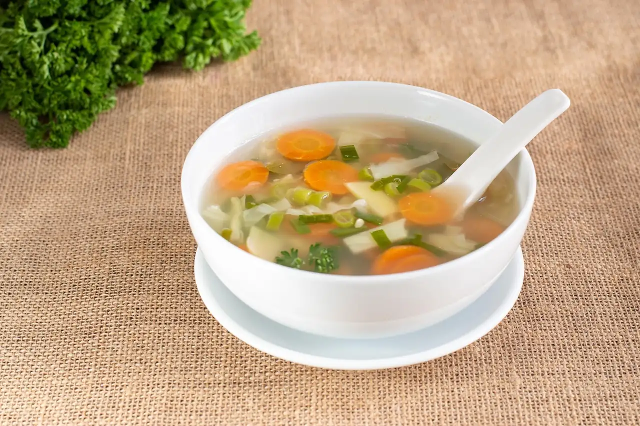 Bouillon clair aux rondelles de carotte, chou et oignon vert dans un bol blanc posé sur une toile de jute