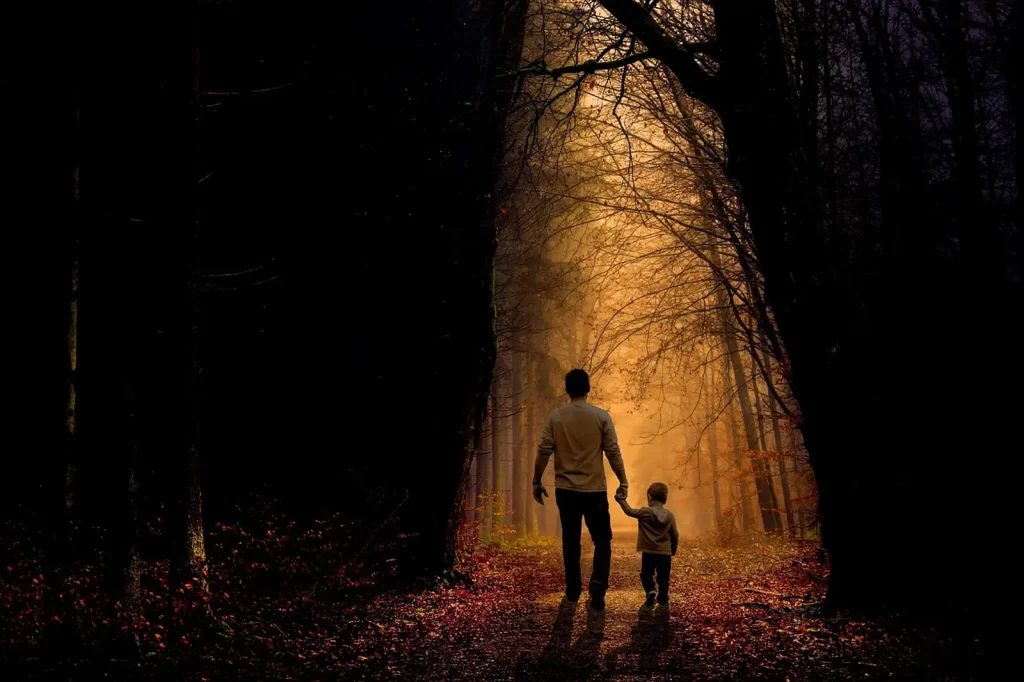 Un père et son jeune enfant marchent main dans la main sur un chemin de forêt couvert de feuilles d’automne