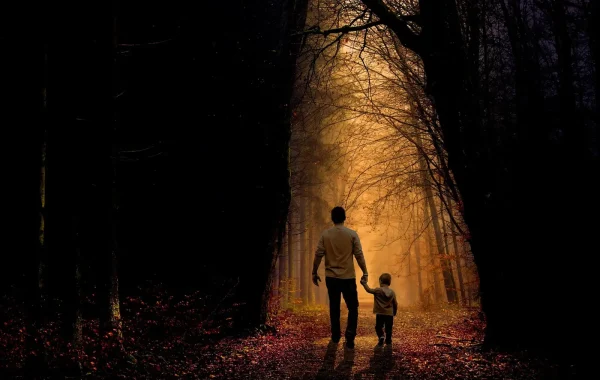 Un père et son jeune enfant marchent main dans la main sur un chemin de forêt couvert de feuilles d’automne