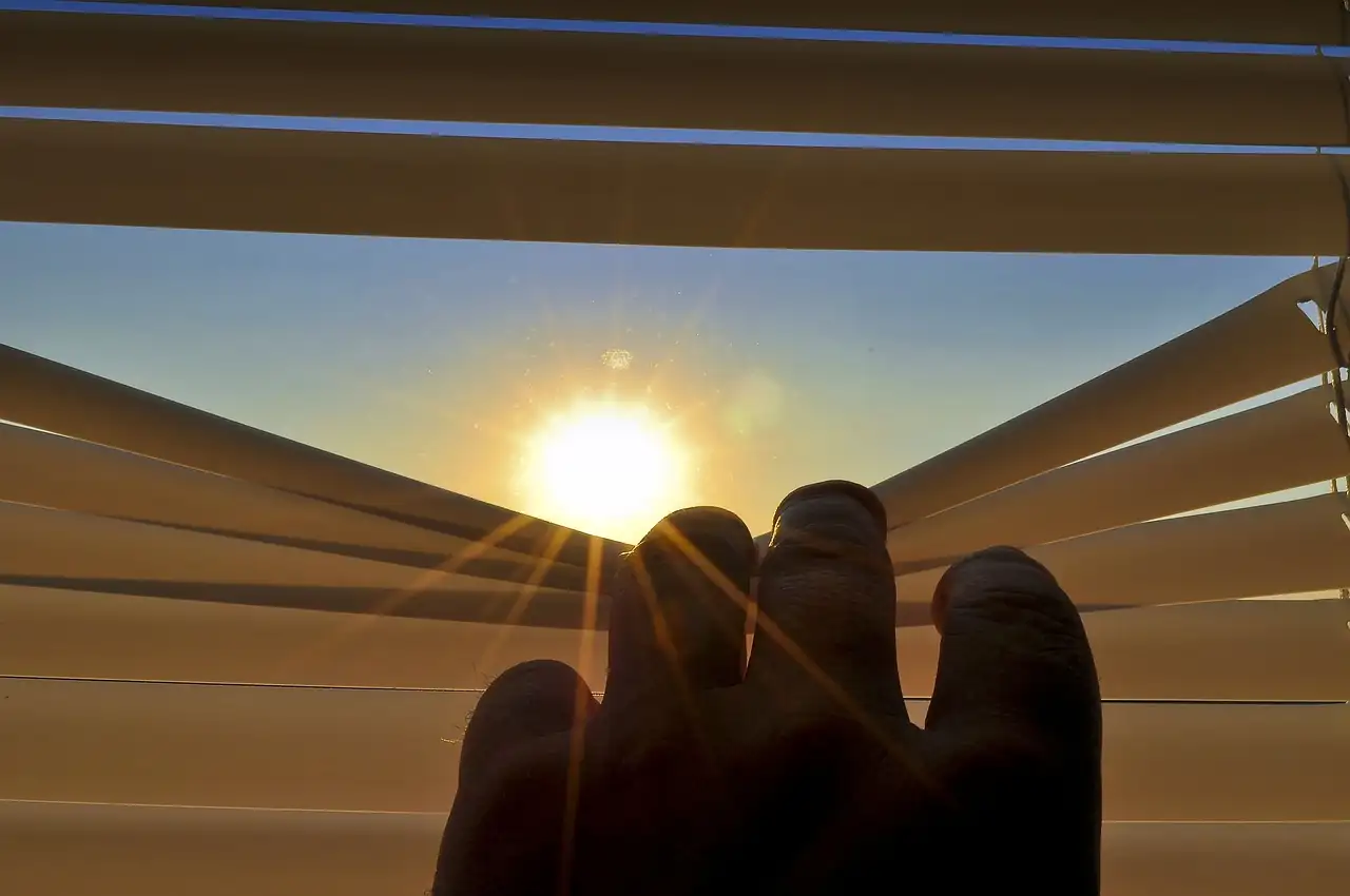 Main qui écarte les lames d’un store devant le soleil levant dans un ciel bleu