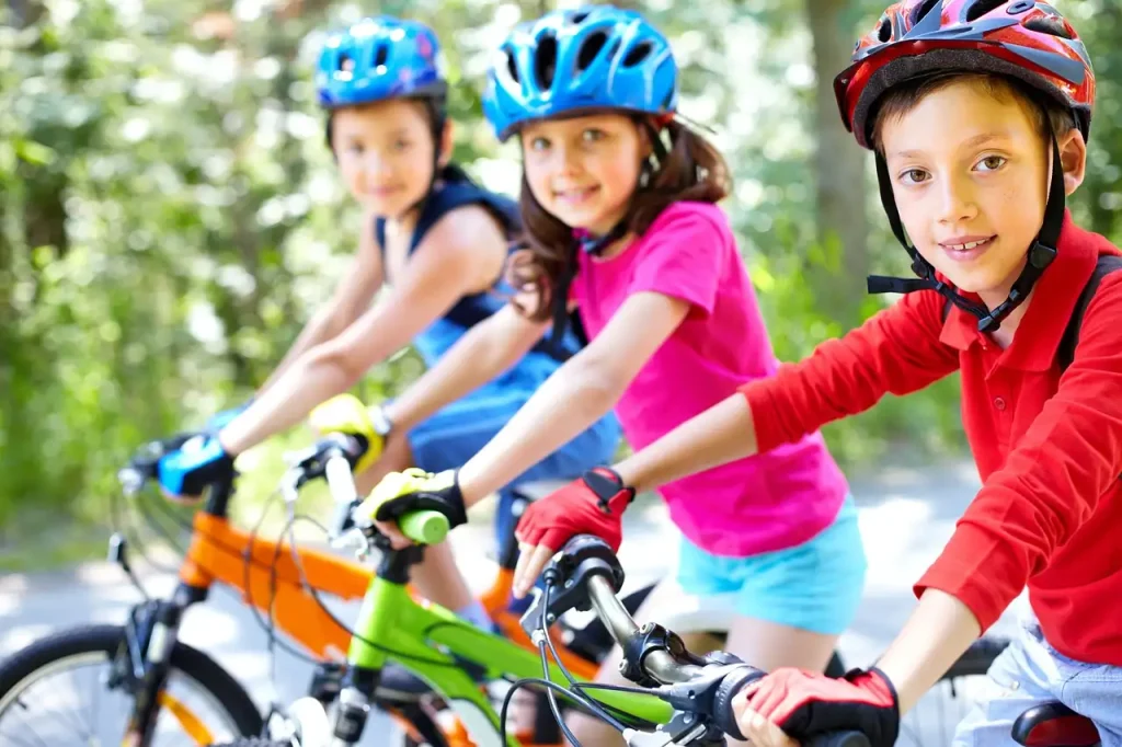 Trois enfants souriants à vélo sur un chemin boisé, casques colorés et gants de cyclisme