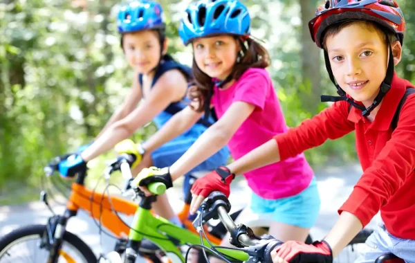 Trois enfants souriants à vélo sur un chemin boisé, casques colorés et gants de cyclisme