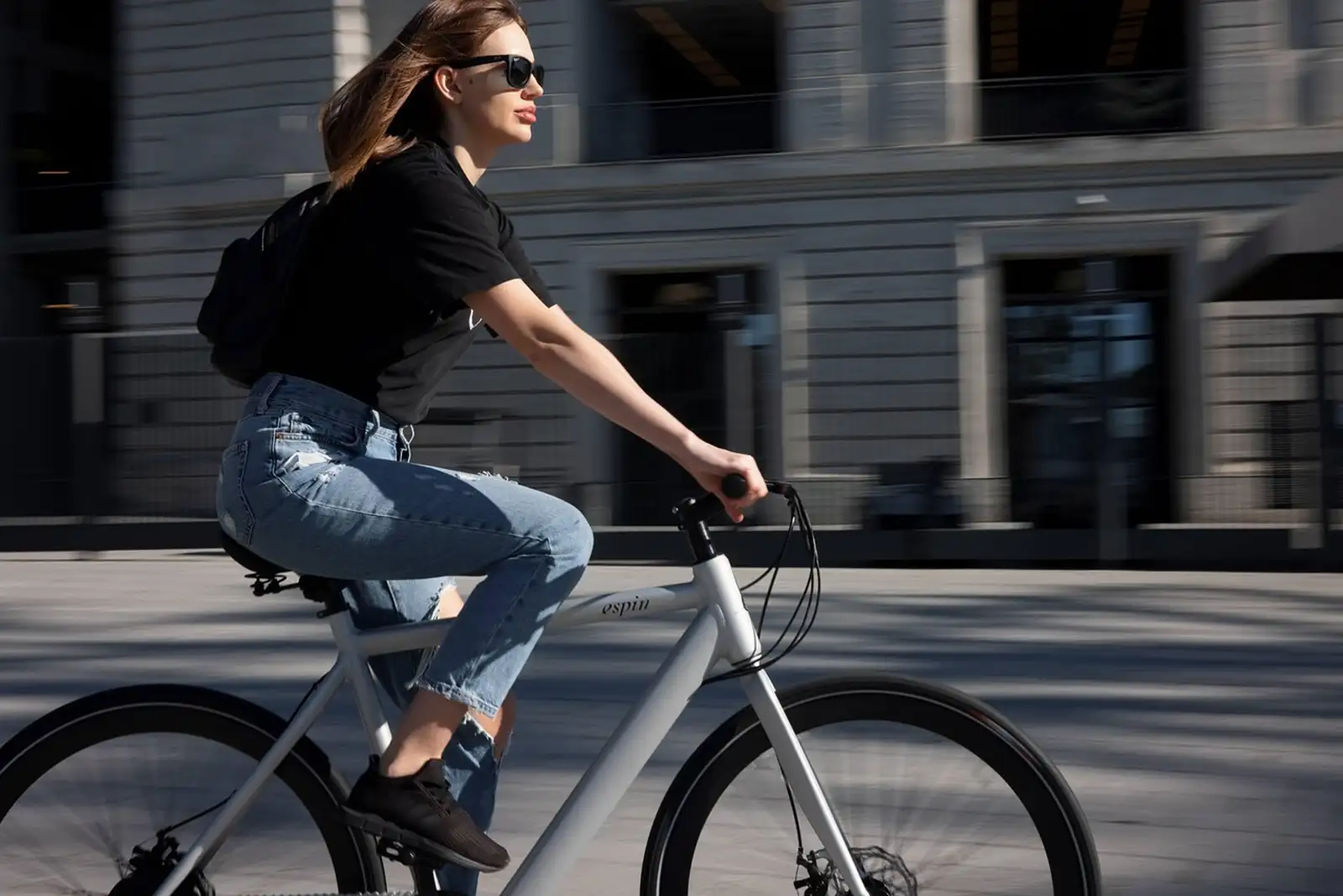 Jeune femme à lunettes de soleil qui roule à vélo dans une rue ensoleillée, devant une façade grise