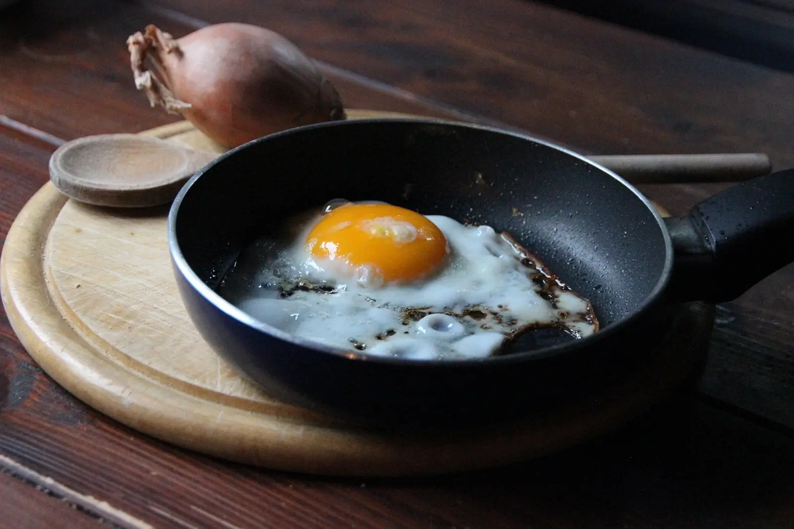 Œuf au plat qui cuit dans une petite poêle noire posée sur une planche en bois, une échalote derrière