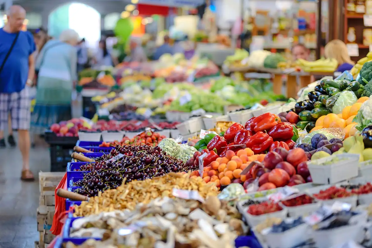 Étals d'un marché couvert chargés de cerises, poivrons rouges, abricots et champignons, clients flous au fond