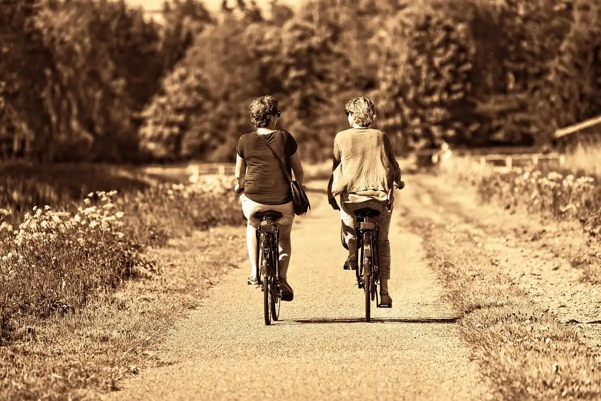 Deux femmes à vélo vues de dos sur un chemin de campagne bordé de fleurs, photo aux tons sépia