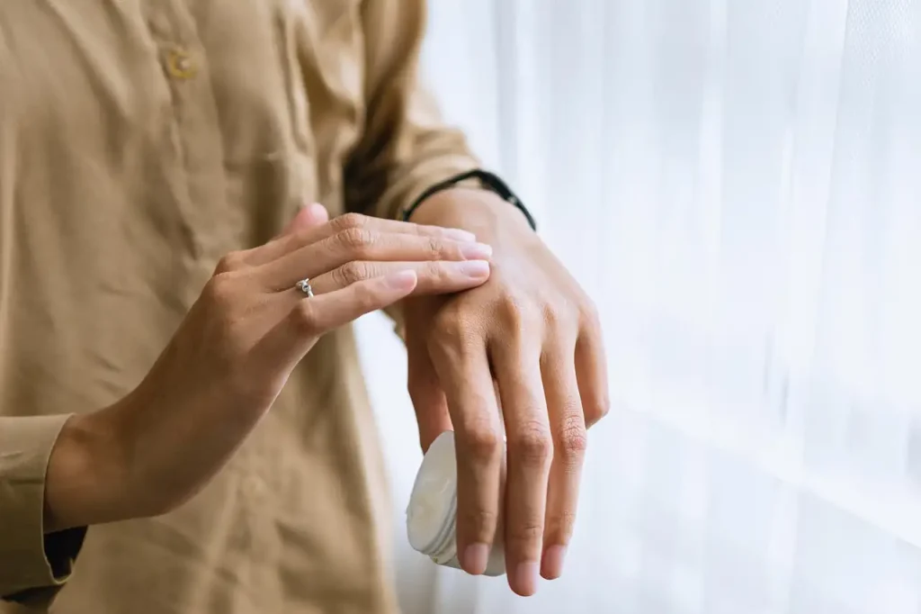 Femme appliquant une crème hydratante sur le dos de sa main, près d'une fenêtre aux voilages blancs